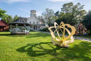 3D Sculpture made of yellow metal on lawn, in front of white gazebo and building